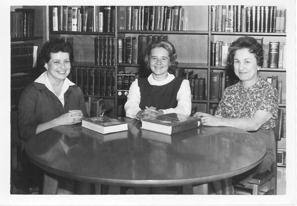 photo of three women around a table in front of book shelves. vintage image black/white.